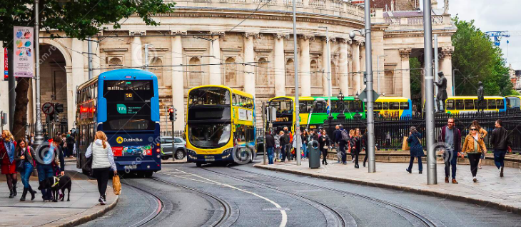 Tram lines and buses in central Dublin and people walking on footpaths. Historic bank building in background